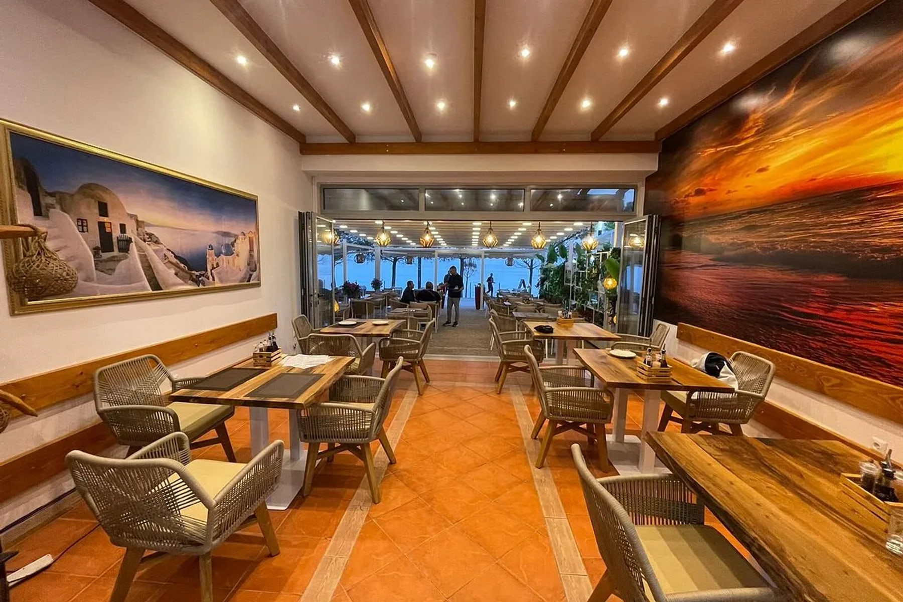 The indoor dining room, terracotta tiled floor and timber-beamed ceiling, opening through folding glass onto a lit terrace and the sea