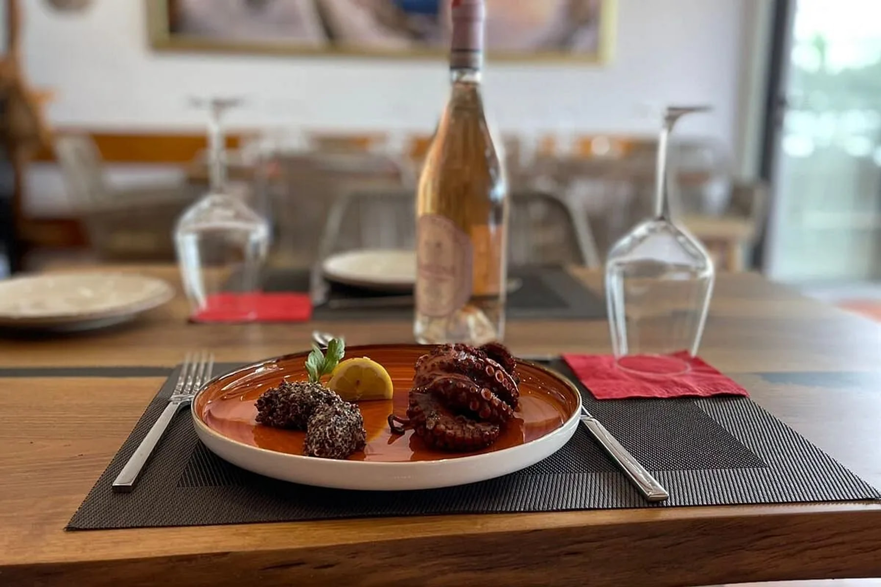 Grilled octopus with lemon and two dark grain croquettes on a copper-glazed plate, a bottle of rosé and upturned glasses behind it on a live-edge timber table