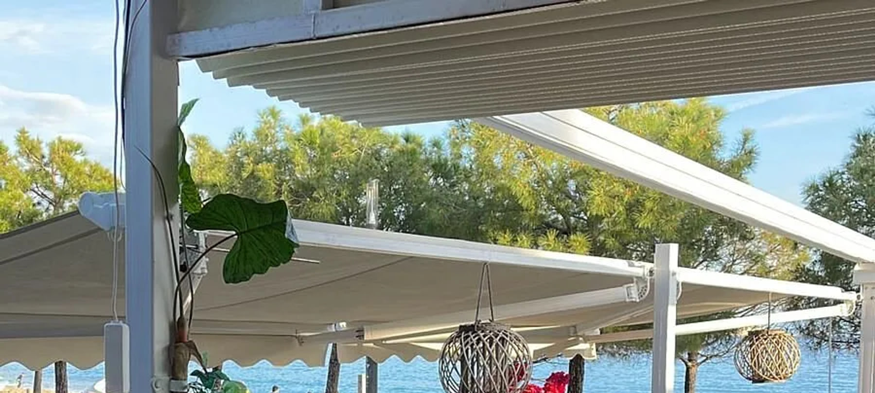 White retractable awnings and a pergola over the terrace, woven rattan lanterns hanging beneath them, pines and the turquoise Ionian behind