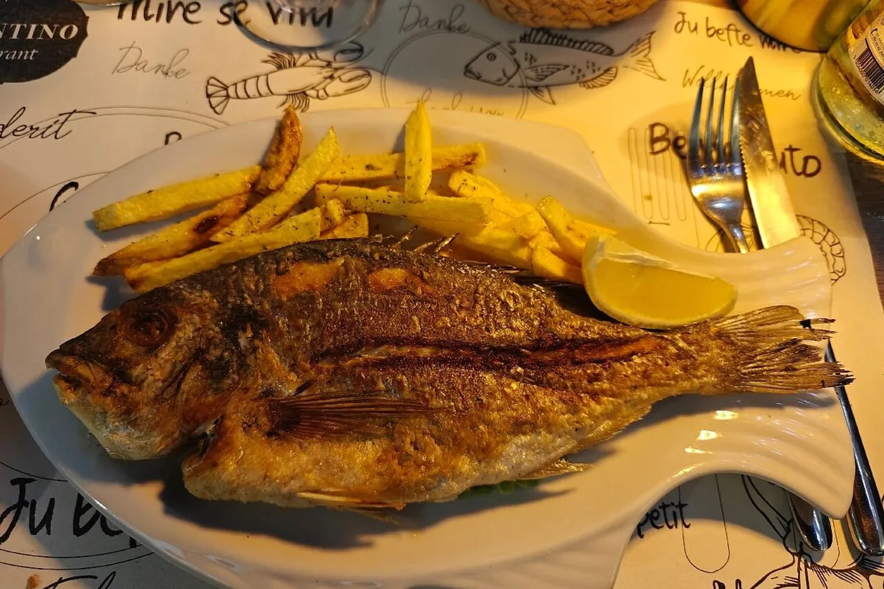A whole grilled fish on a fish-shaped white plate with chips and a wedge of lemon, on a placemat printed with the Valentino Restaurant mark