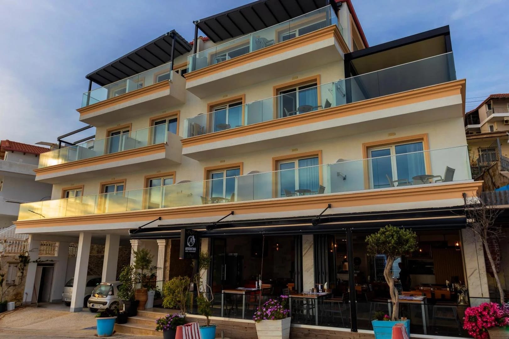 A four-storey cream building with ochre window surrounds and glass balconies, its ground-floor restaurant open to the street at golden hour