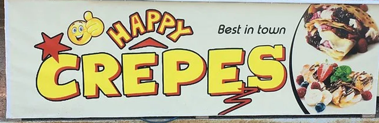 The printed banner along the front of the counter: HAPPY CRÊPES in fat yellow letters outlined in red on an off-white vinyl ground, with a cartoon smiley, the words Best in town, and two photographs of filled crepes at the right.