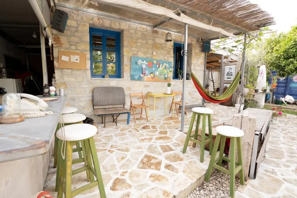 Flagstone courtyard at Himara Hostel with green bar stools, a yellow folding table, a hammock and a reed sun-shade against the stone house