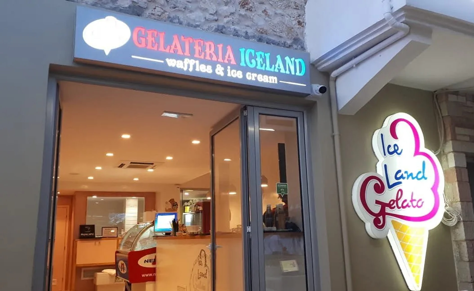 The lit shopfront of Gelateria Iceland in Spile, Himarë, its fascia reading GELATERIA ICELAND above an open bi-fold door