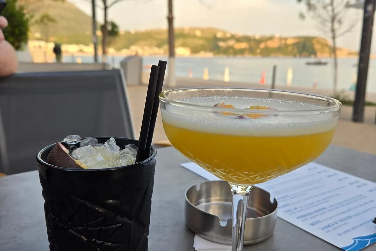 A foam-topped coupe of deep amber cocktail and a black tumbler of crushed ice on a promenade table, with Himarë bay and a wooded headland behind them in low sun