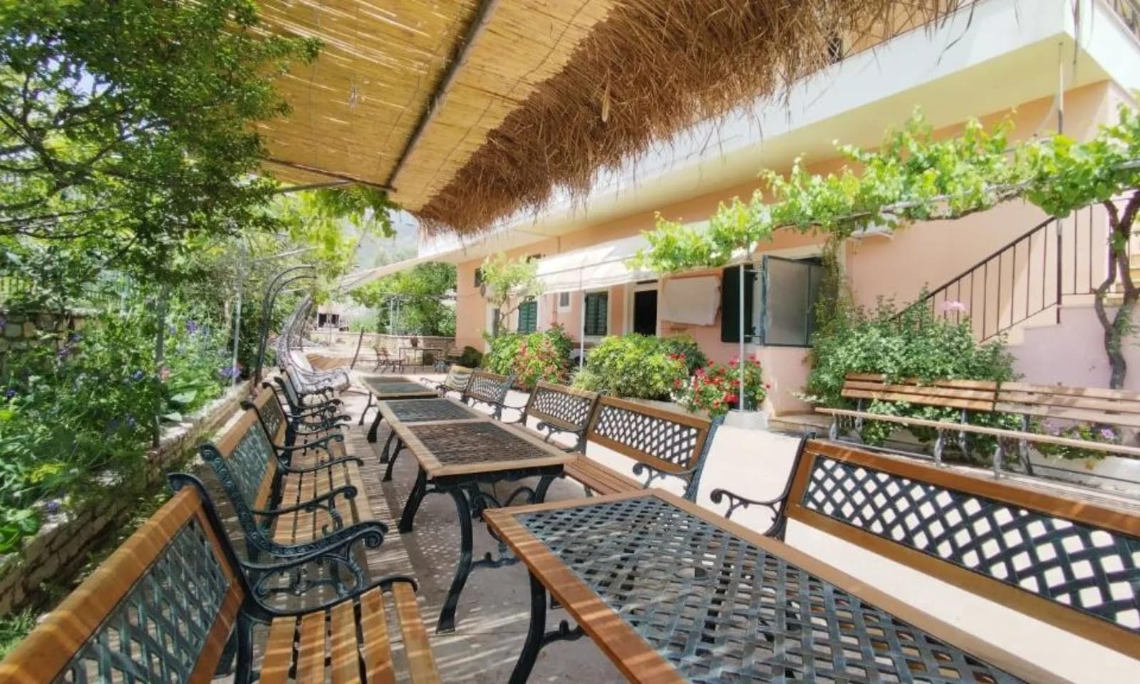 Benches and long tables under a reed pergola beside the apricot-painted wall of Oasis Hostel in Himarë