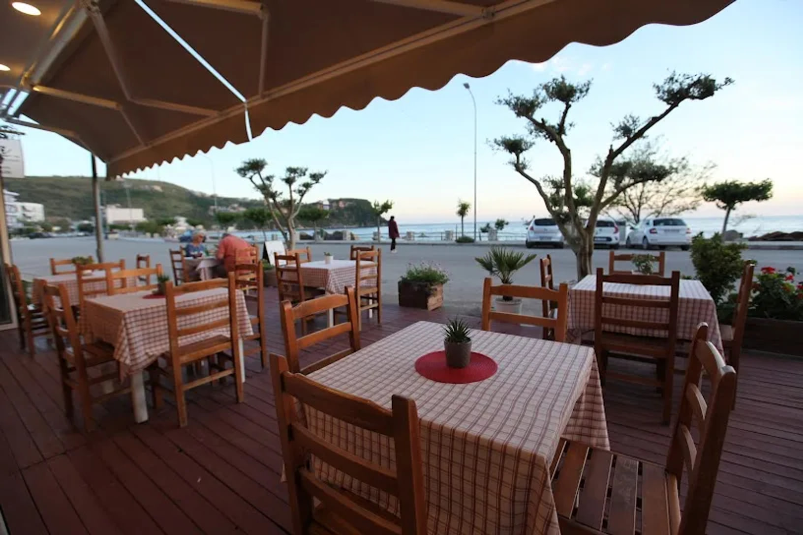 Wooden tables under a brown scalloped awning on the terrace at Restaurant Kolagji, the road, the promenade and the sea beyond at dusk