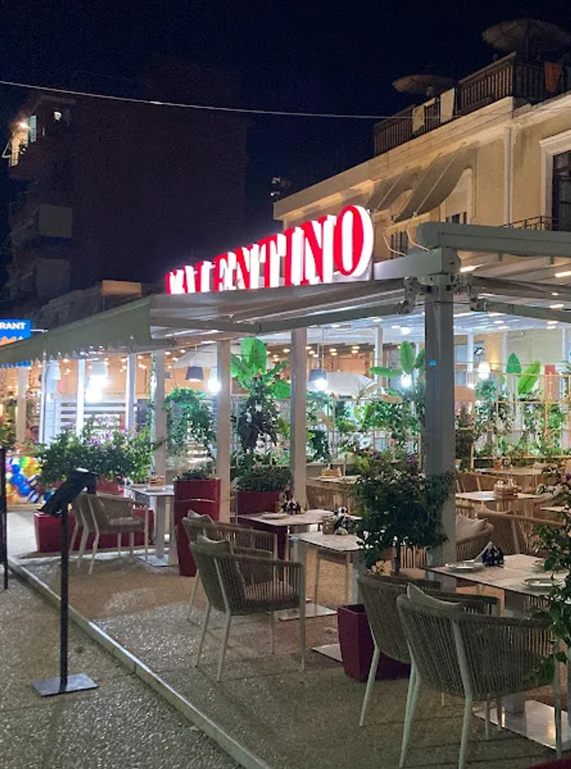The red neon VALENTINO sign lit above a white pergola on the Himarë seafront at night, with set tables and rope-weave armchairs beneath it