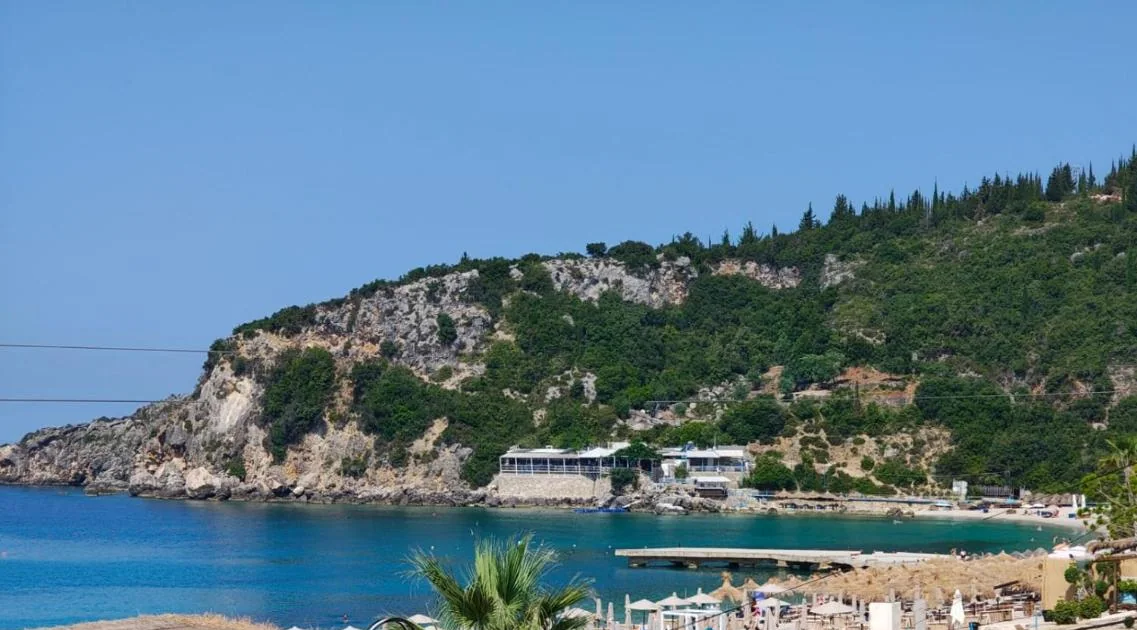 Livadhi Bay from the property: the pine-topped headland, the jetty and rows of straw parasols along the shingle.
