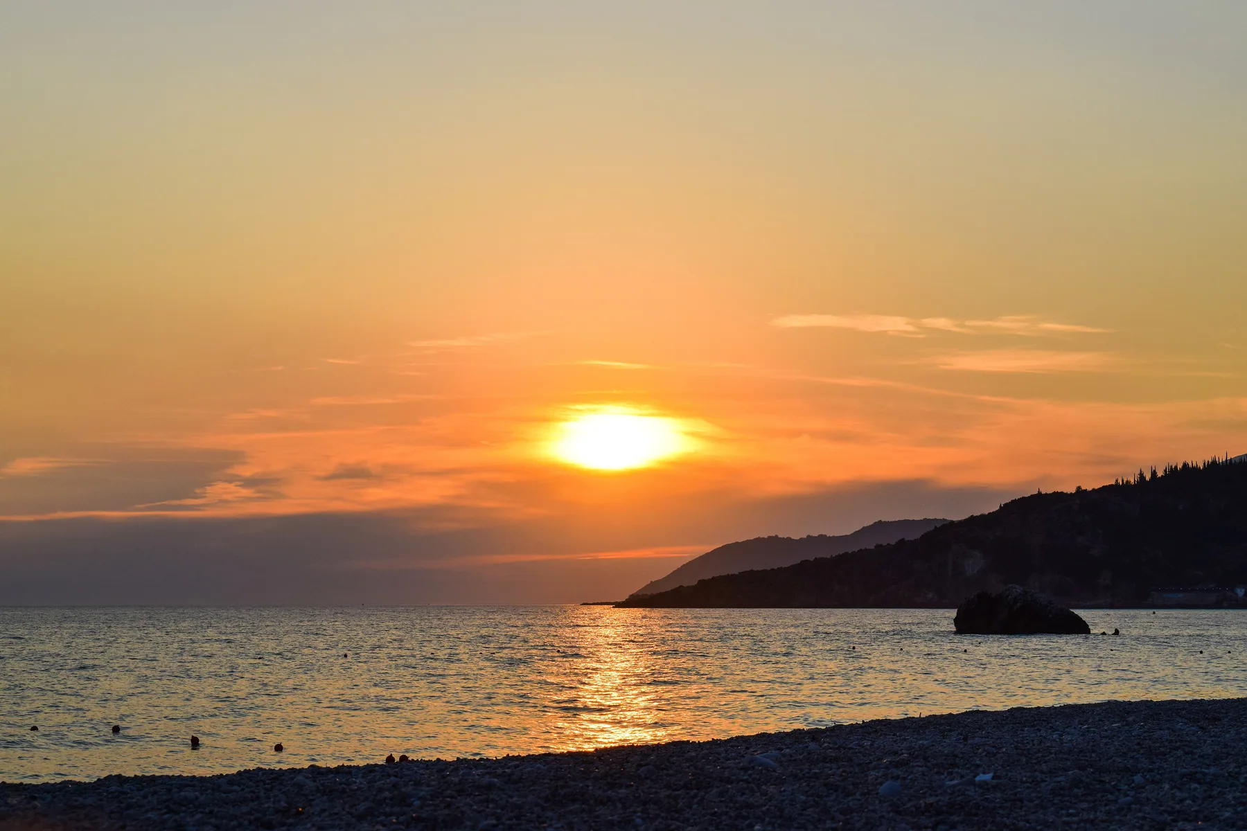 Livadhi Beach at dusk: the sun going down over open water, the headland off to one side, the shingle in the foreground.