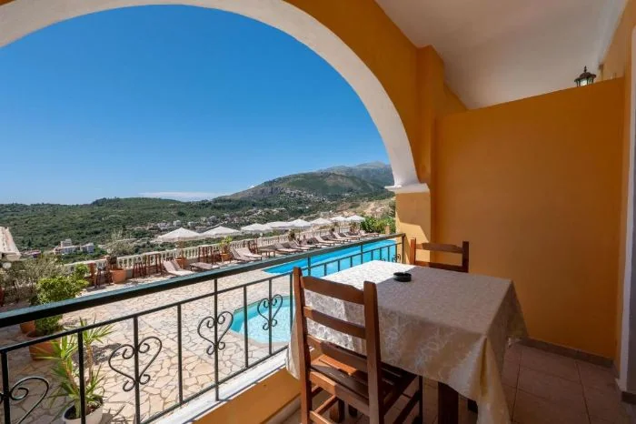 An arched balcony at Apart Hotel Alex opening onto a table and chairs, the pool below and the hills across the valley