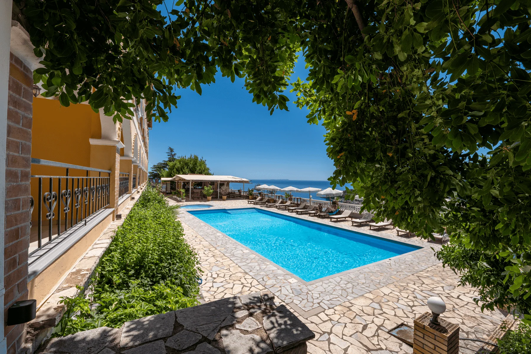 The pool at Apart Hotel Alex seen from under an overhanging tree, the yellow arcade to the left and the Ionian beyond the deck