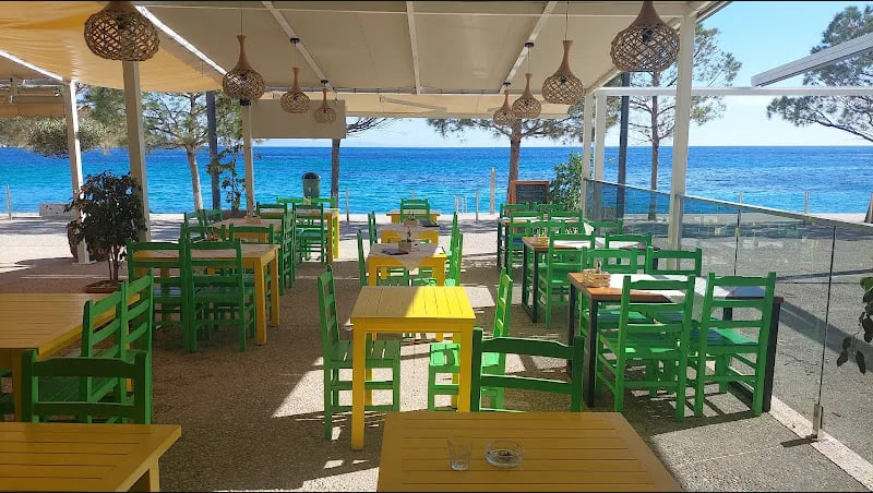 Green and yellow painted tables and chairs on exposed-aggregate concrete under a cream stretched awning, the sea beyond a glass balustrade