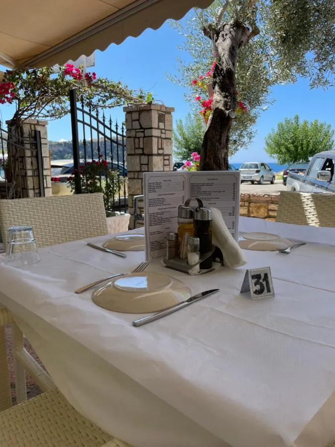 A table laid with white linen under a cream awning on the restaurant terrace, an olive tree growing through it, bougainvillea over the stone gate posts and the sea beyond