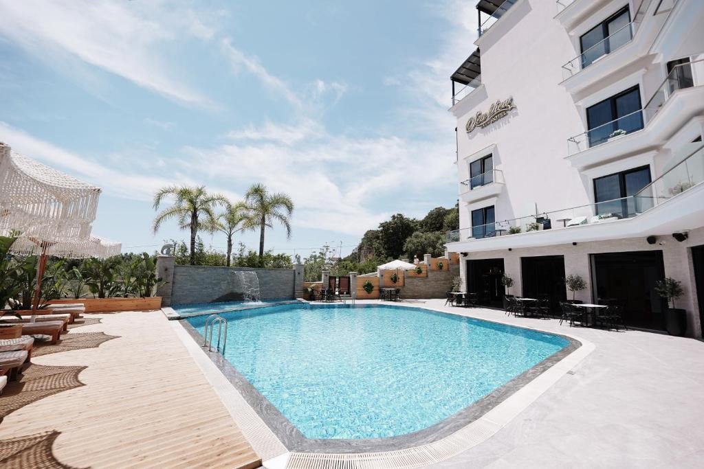 The hotel pool from the timber deck, with sun loungers and fringed parasols on the left, palms behind, and the white hotel and its ground-floor terrace tables on the right