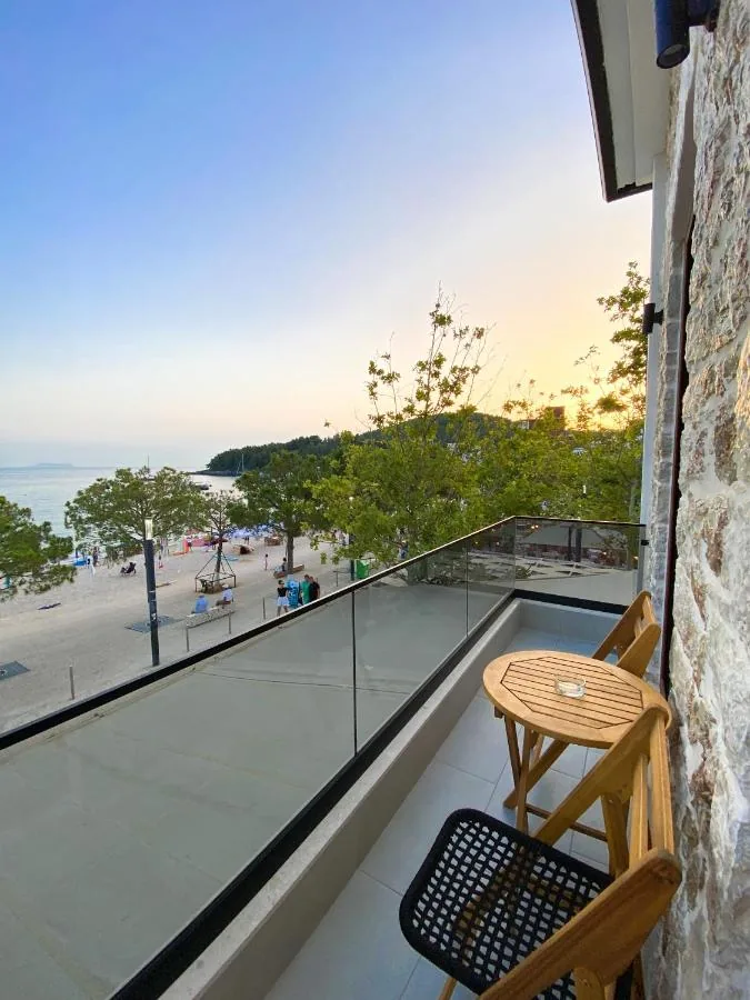 A balcony with a glass balustrade and a folding timber table at dusk, looking down onto the paved beachfront street, the sand, the moored boats and the headland with the last of the sunset behind it.