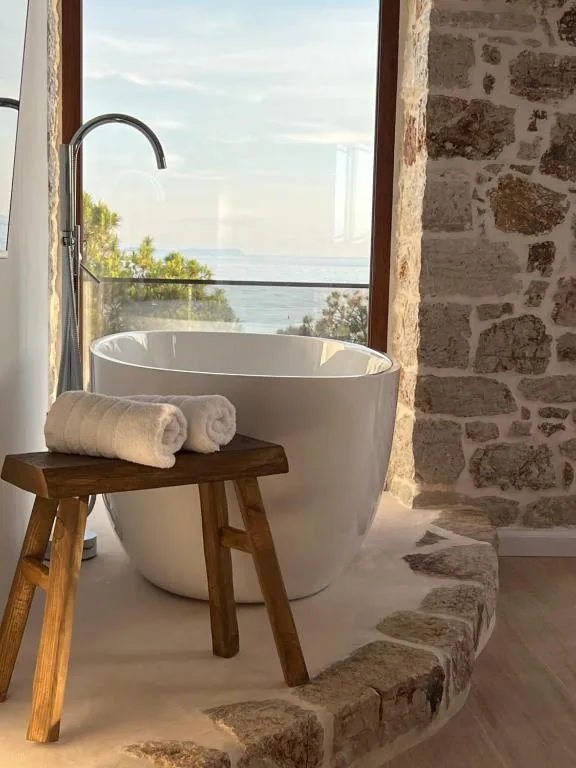 A freestanding bath seen past a rough timber stool carrying two rolled white towels, with the sea and pines through a full-height glazed door and rubble limestone to the right.