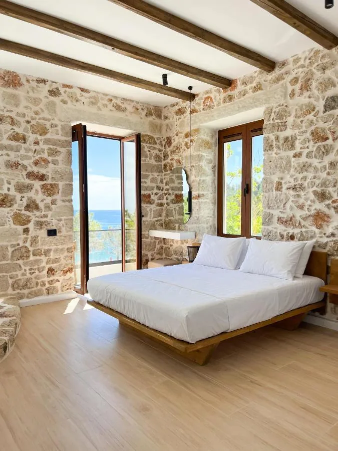 A wide bed on a low timber platform, rubble limestone on every wall in the frame, a glazed door standing open onto a balcony above the sea, and a two-leaf window onto green.