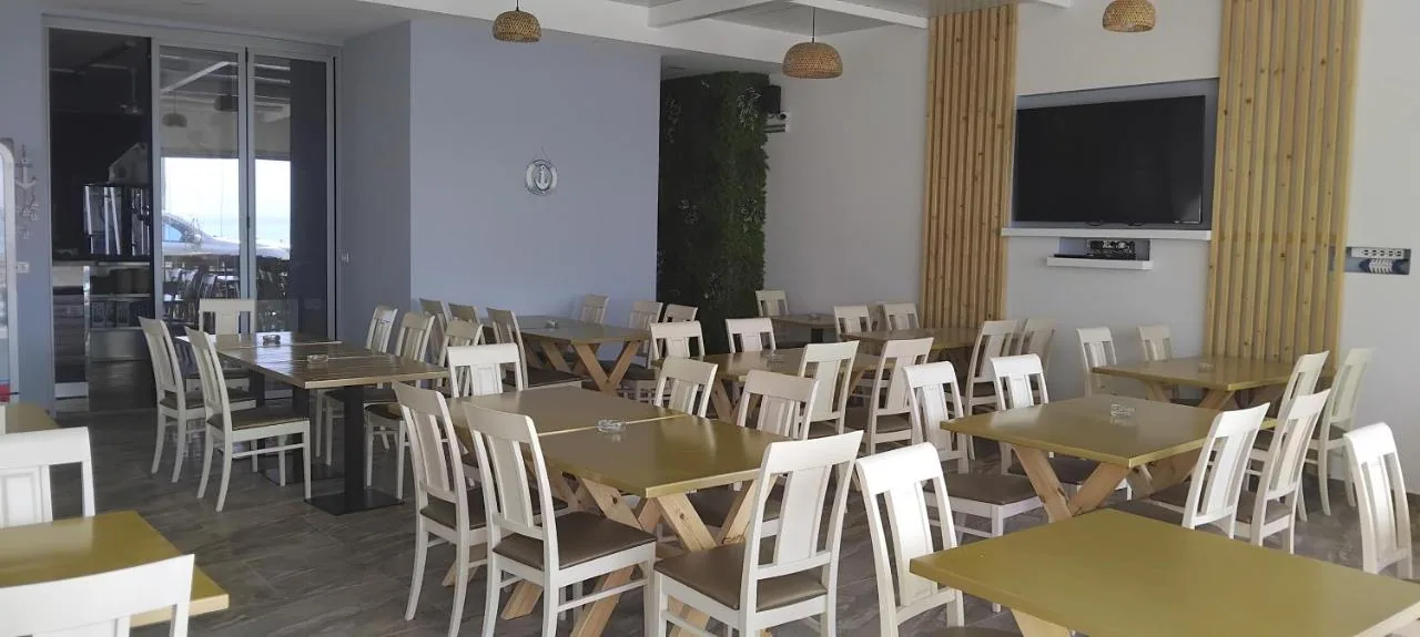 Bare timber tables in the dining room at Anxhelos, with the preserved-moss wall, the oak fins and the covered terrace through the sliding doors.