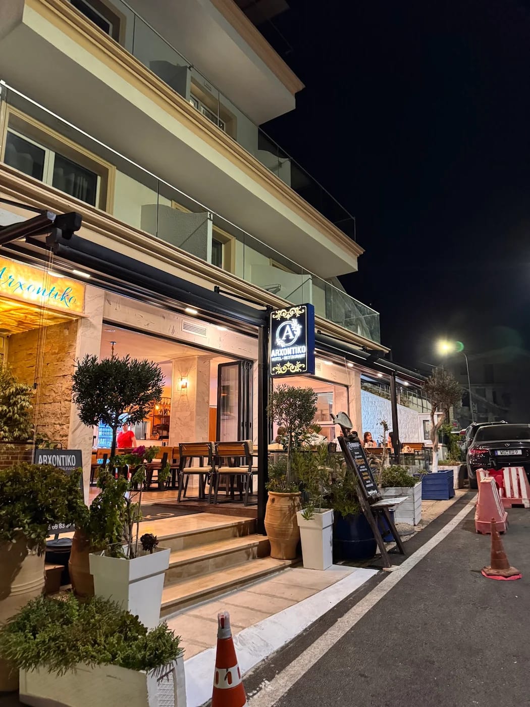The lit frontage of the restaurant at night, the ARXONTIKO sign lettered in gold on its dark blue plaque beside the pavement tables