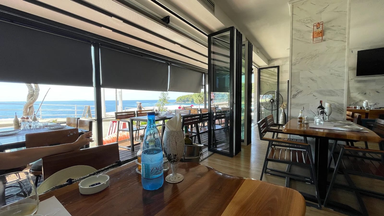 The dining room with marble walls and live-edge timber tables, its folding glass front open to the terrace and the sea