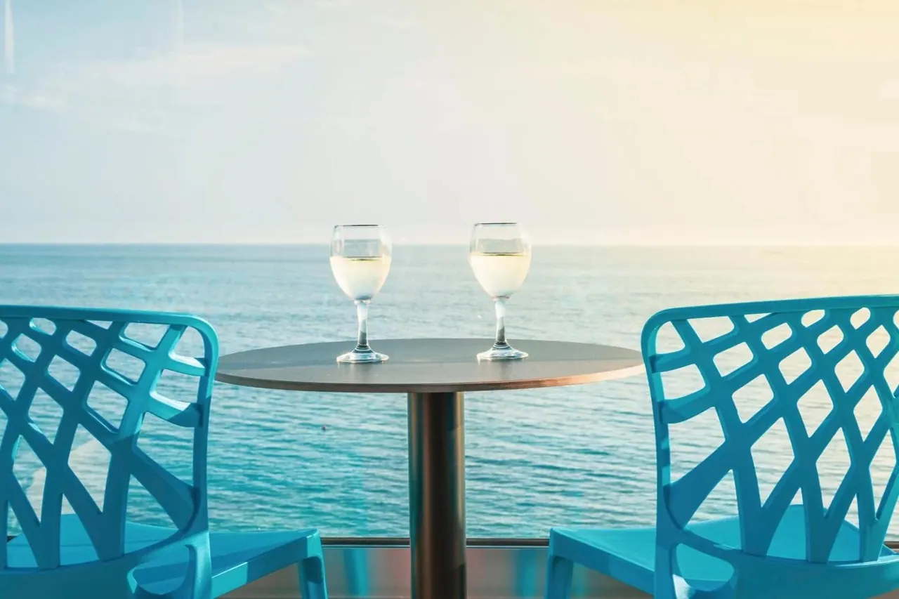 Two glasses of white wine on a balcony table between turquoise chairs, with the sea behind