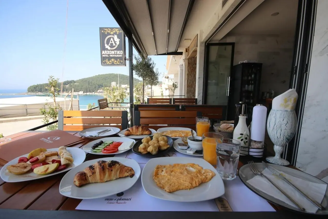 A breakfast table on the terrace with the black-and-gold ARXONTIKO plaque above it and Himarë bay behind