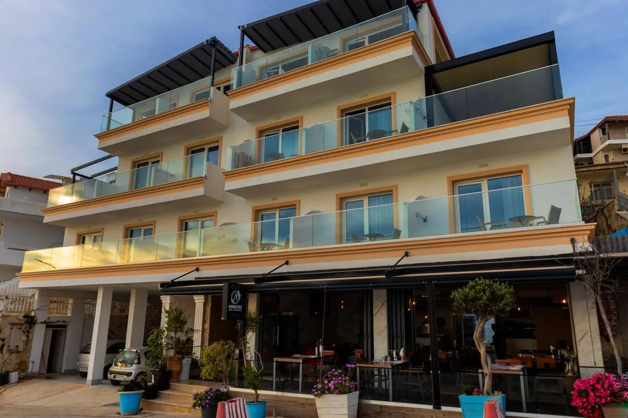 A four-storey cream building with ochre window surrounds and glass balconies, its ground-floor restaurant open to the street at golden hour