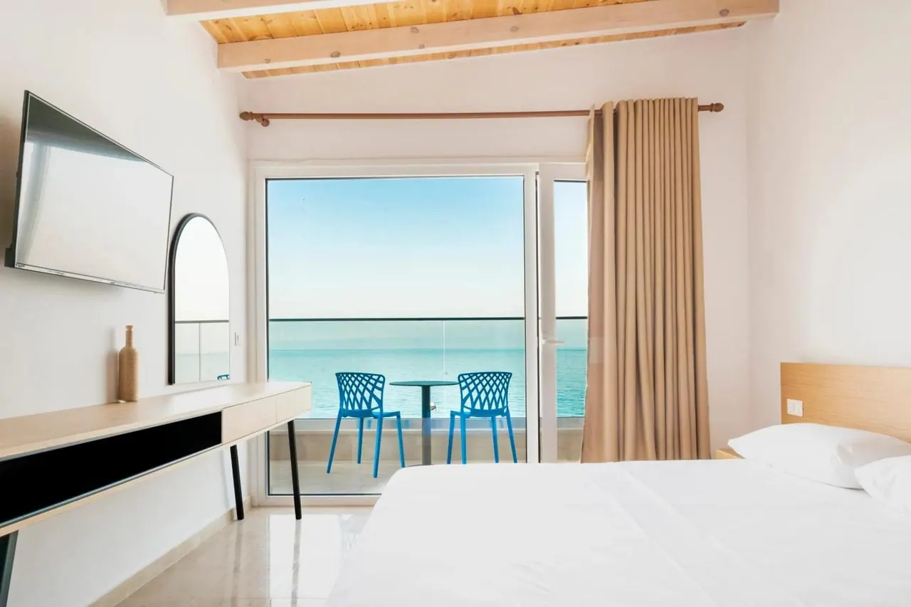 A white-linen bed beside a glazed balcony door, with two blue chairs, a table and the sea beyond
