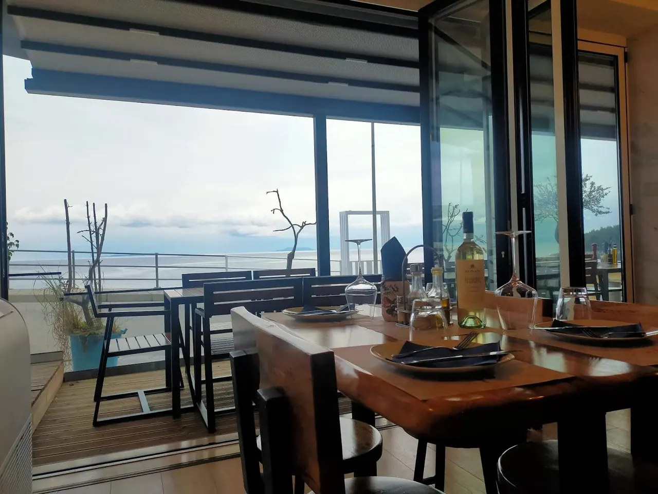 A laid table inside the restaurant, black plates and a bottle of white wine, with the deck and the open sea beyond the folded-back frontage