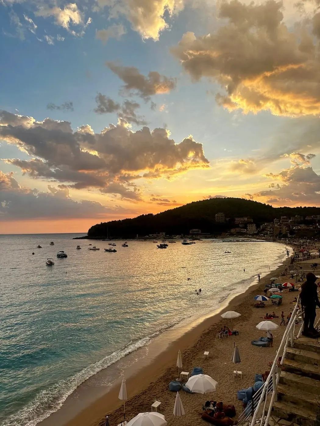 Himarë bay at dusk from above the beach, with moored boats, umbrellas on the sand and the pine headland