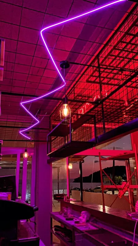 A violet neon zigzag across the terrace ceiling at night, with a caged filament lamp and the dark sea beyond