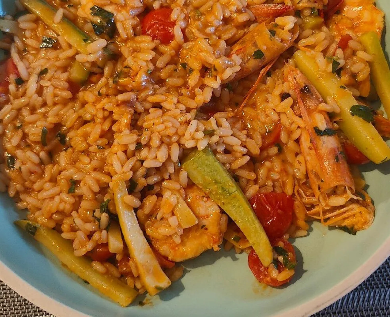 Close view of tomato-tinted rice with whole prawns, cherry tomatoes, courgette batons and chopped parsley, served in a pale celadon bowl