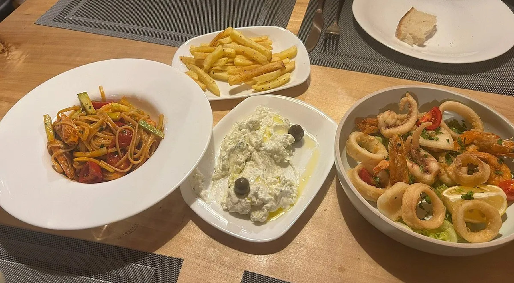 Four dishes across a timber table — spaghetti with prawns, cherry tomato and courgette; chips; a lumpy white dip under olive oil with two black olives; and a bowl of squid rings, tentacles and whole prawns with lemon — beside a side plate holding a torn piece of bread