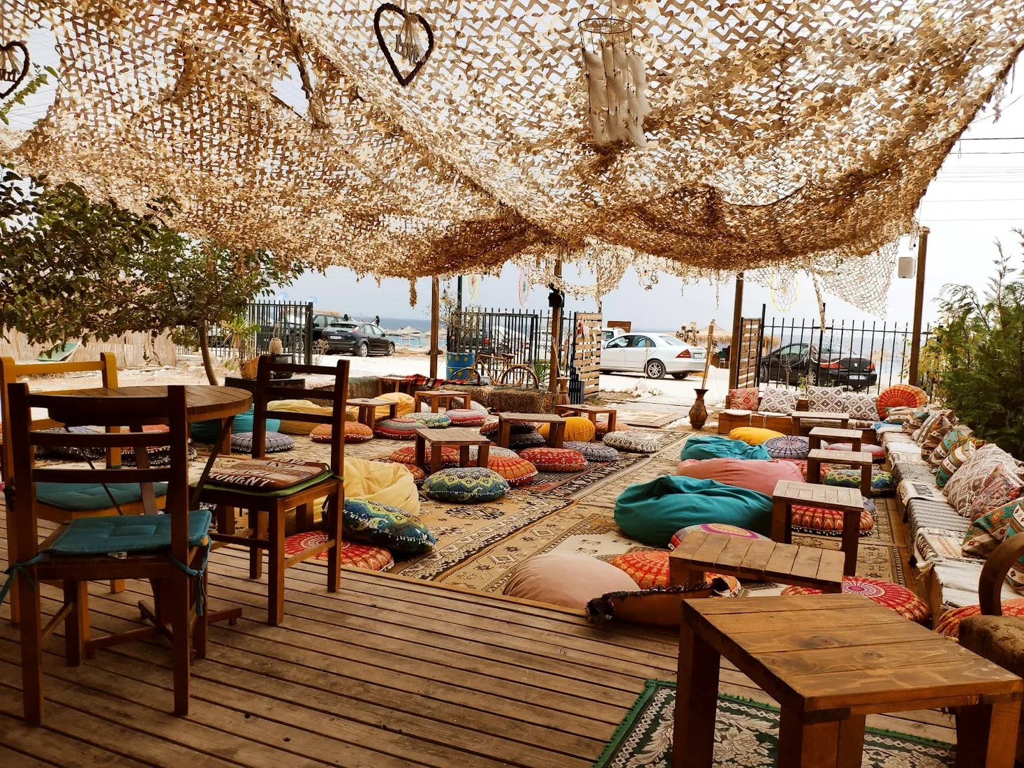 Floor cushions and layered kilims under a wide canopy of pale camouflage netting at Boho Livadh, with the parking strip and the sea beyond