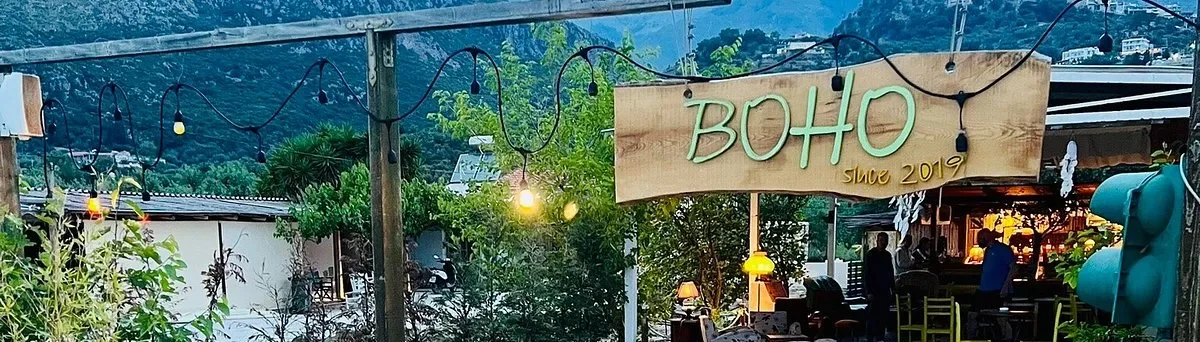 Boho Livadh's hand-painted timber sign reading BOHO since 2019, a festoon string crossing it, against the Himarë hills