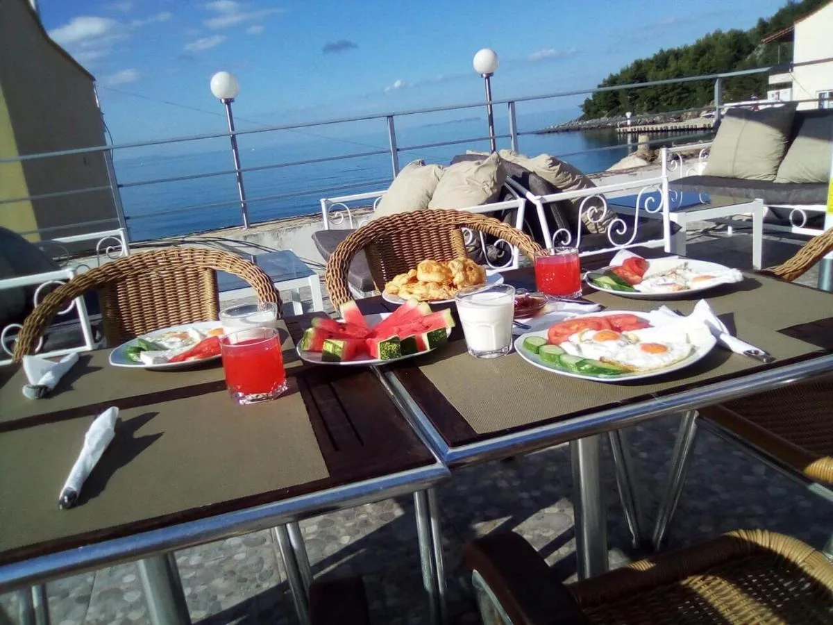 Breakfast laid on the roof terrace in morning sun — fried eggs, tomato, cucumber, white cheese, watermelon and juice, with the bay behind.