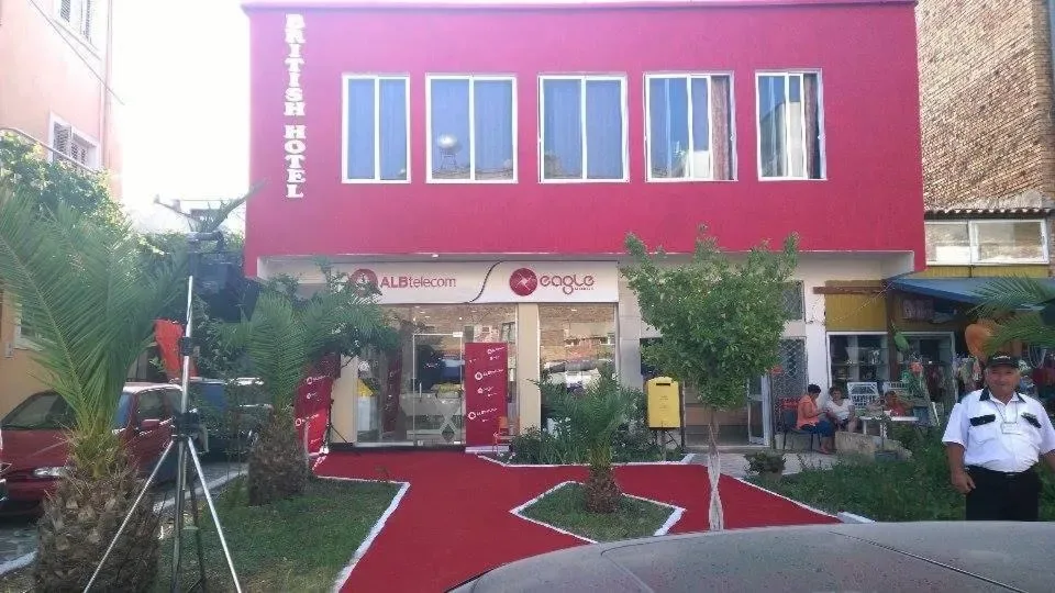 The red painted street elevation of British Hotel in Himarë, its name lettered vertically down the corner above ground-floor shops.