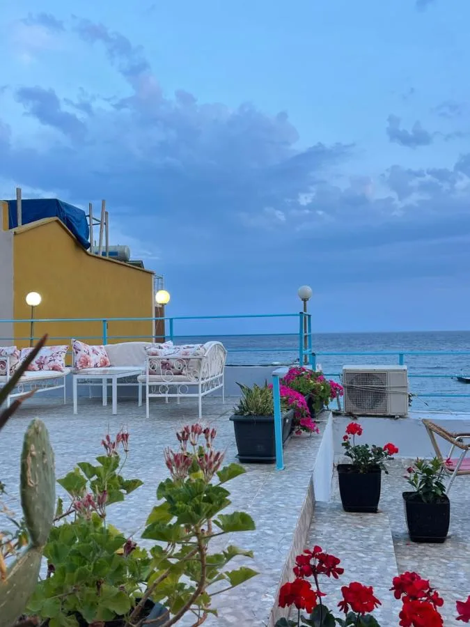 The hotel roof terrace at dusk, with turquoise railings, white seating, potted geraniums and the open sea beyond.