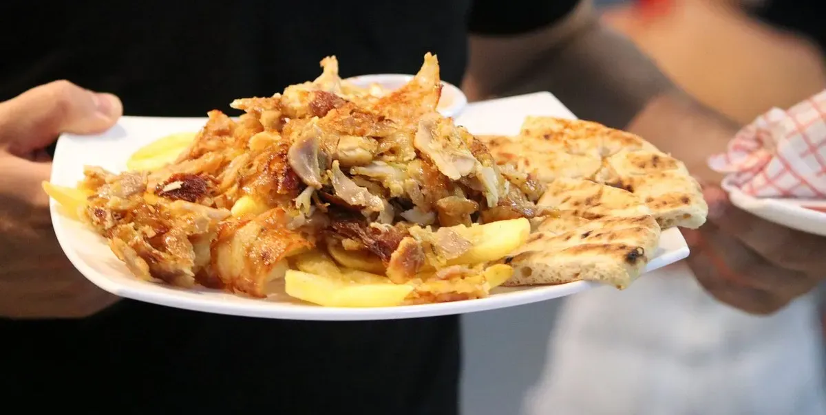 A white plate of carved meat over chips with grilled flatbread and a small pot of sauce, held in a hand, with a second plate lined with red-checked paper behind it