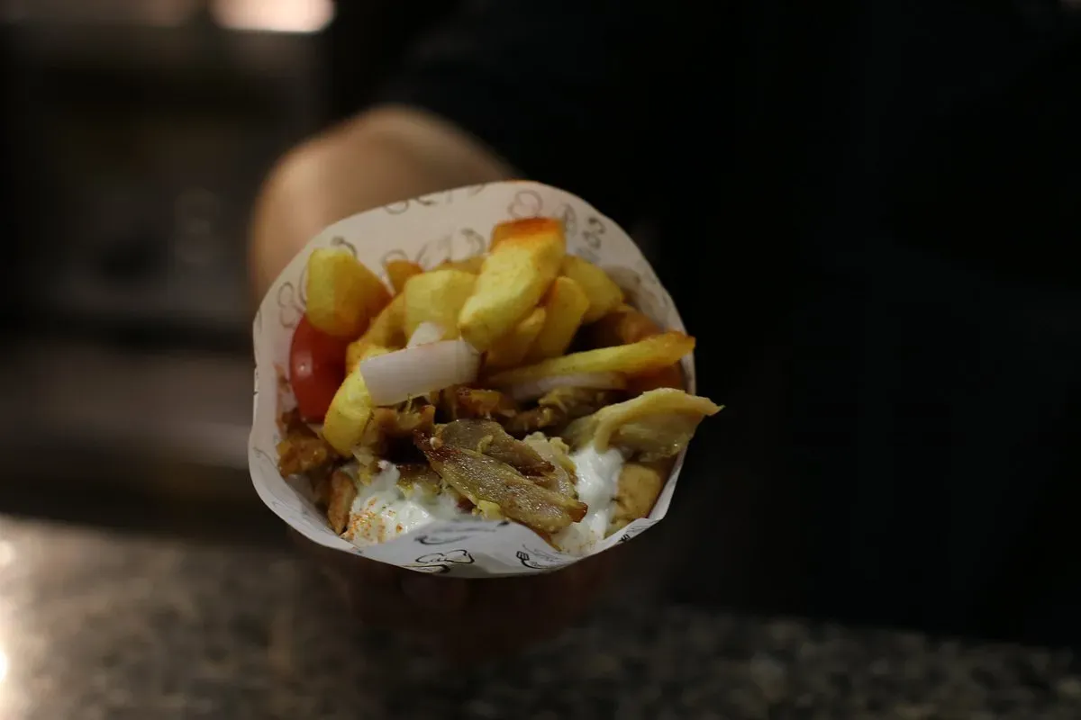 A cone of printed paper held in one hand over a stone counter, filled with chips, carved meat, a whole cherry tomato, raw onion and a white sauce