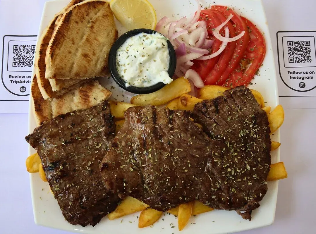 A white plate of three grilled cuts of meat over chips with grilled flatbread, a lemon wedge, sliced tomato, sliced red onion and a pot of white sauce, on a placemat printed with the venue's TripAdvisor and Instagram QR codes