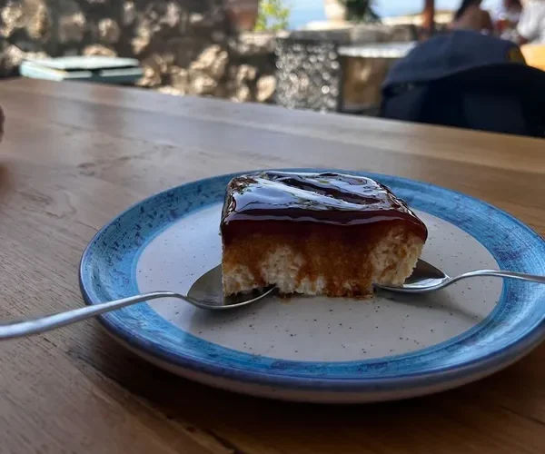 A square of caramel-topped milk cake on a blue-rimmed speckled plate with two spoons, on a bare timber table, the stone parapet and the terrace out of focus behind