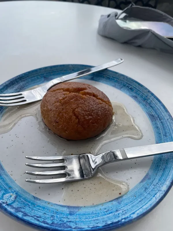A round golden syrup-soaked cake in a pale sauce on a blue-rimmed speckled plate, a fork and a knife laid on either side of it, on a white round table