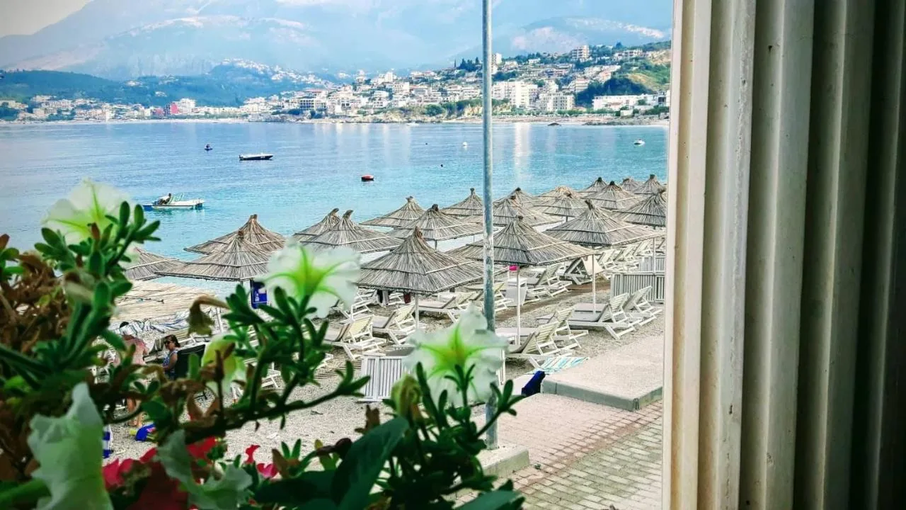 The Himarë bay from the colonnaded terrace at Castle Hotel: thatched parasols and white loungers on the beach, boats on flat turquoise water, the town and its hills across the bay