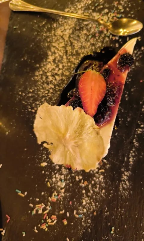 A wedge of pale set dessert under dark berry compote and a whole blackberry, with a halved strawberry and a large dried pineapple slice, on dark slate strewn with sugar and coloured sprinkles beside a gold spoon