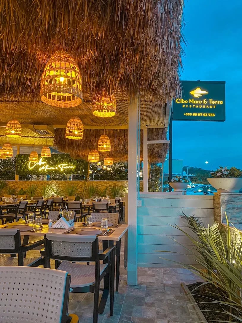 The terrace of Cibo Mare e Terra at dusk: laid tables under a thatched roof hung with lit rattan lamps, the restaurant's illuminated sign at the right against a blue evening sky