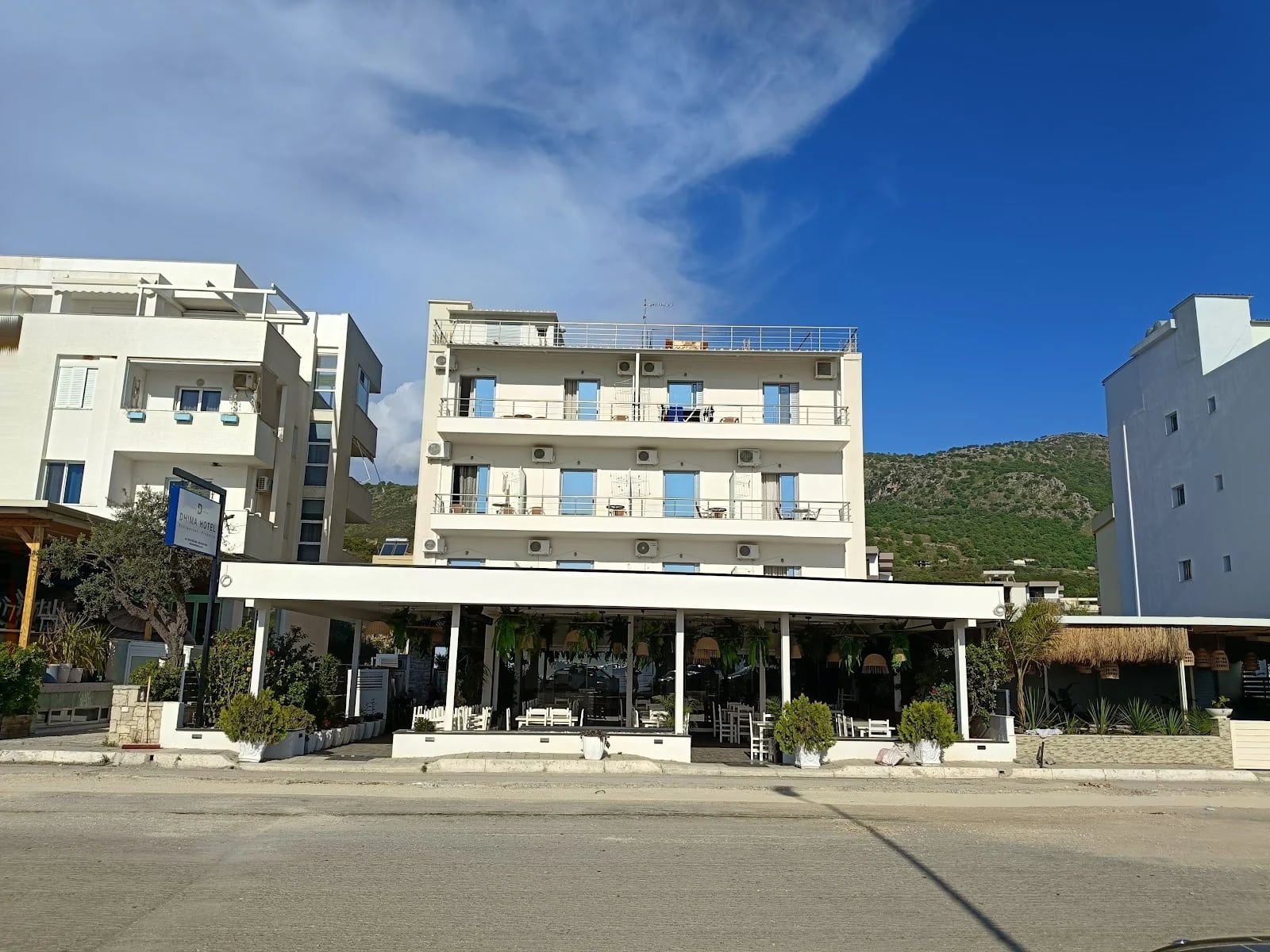 The four-storey white Dhima Hotel, three floors of balconies over a restaurant terrace under a flat canopy, seen from the coast road