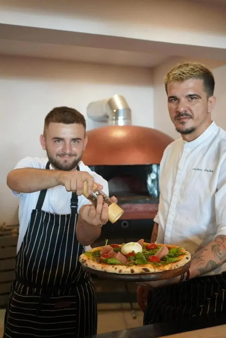 Two chefs in aprons in front of a copper-domed pizza oven, one holding a finished pizza