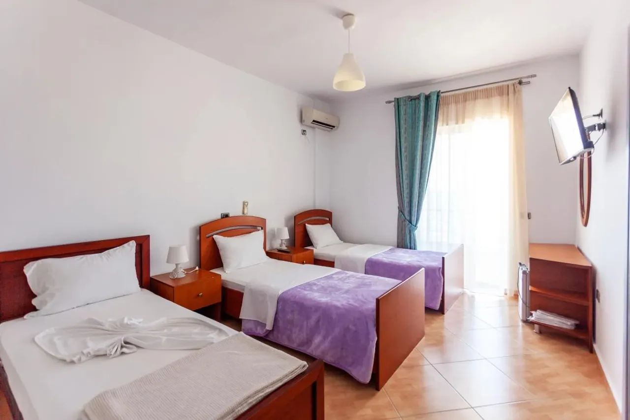 Three single beds under purple throws, teal curtains at a full-height window and an air-conditioning unit above